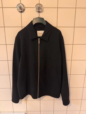 Svart jacka från Les Deux - Stilren svart jacka från Les Deux med rak passform och klassisk krage. Jackan har dragkedja framtill och två sidofickor. Materialet är mjukt och ger en clean look som passar till många olika outfits.