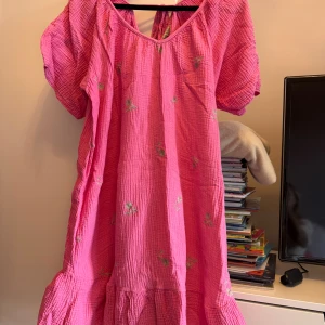 Rosa strandklänning Made in Italy - Oversize klänning med broderade blommor och volang nertill. Klänningen har puffiga korta ärmar, knytning i ryggen och är gjord i ett mjukt, krinklat bomullstyg (muslin). Känningen är one size men passar bra till en person som bär storlek S. 