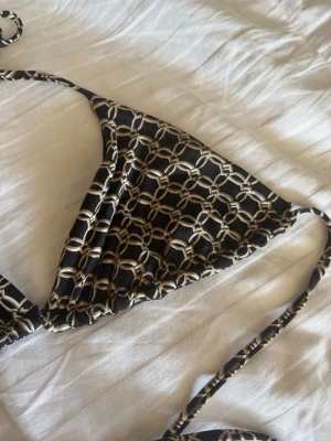 Svart bikiniöverdel med kedjemönster - Snygg trekantsbikini i svart med beige kedjemönster och knytband i nacken och ryggen. Perfekt för stranden eller poolen, enkel att justera för bästa passform. Stilren och trendig design som sticker ut.