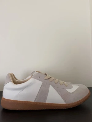 Maison margiela - Stilrena sneakers i vitt och beige med ovandel i mocka och läder. Beige snörning och detaljer, samt klassisk brun gummisula för en retro känsla. Perfekt för dig som gillar minimalistisk och clean stil.