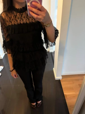 Svart spetsblus med volanger - Trendig svart blus i spets med volangdetaljer och hög hals. Blusen är genombruten och har trekvartsärmar, vilket ger en cool och elegant look. Perfekt för dig som vill sticka ut med en unik topp.