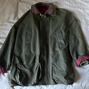 Holland & Holland vintage vaxad jacka - Storlek L. Vintage Holland & Holland wax jacket / jaktjacka – fasanfoder – Made in England-era Säljer en fantastisk vintage waxed cotton-jacka från Holland & Holland, troligen från sent 80-tal eller tidigt 90-tal. Klassisk brittisk shooting/field jacket i mörkgrön vaxad bomull med brun manchesterkrage och ovanligt vinrött foder med fasanmotiv. Riktig “old money / countrywear”-känsla