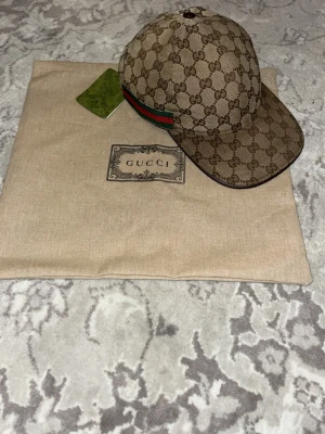 Gucci keps med monogram och röd-grön rand - Snygg keps från Gucci i beige med klassiskt GG-monogram över hela kepsen. Kepsen har en böjd skärm och en ikonisk röd-grön rand på sidan. Materialet är canvas och kepsen levereras med originalpåse och tagg. 