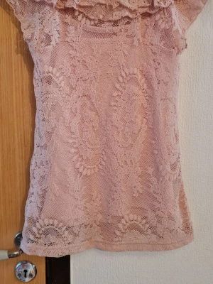 Rosa spetsblus med volang KappAhl - Söt ljusrosa blus från KappAhl i genombruten spets med romantisk volang runt halsringningen. Blusen är ärmlös och har ett feminint, figurnära snitt. Perfekt för dig som gillar en mjuk och elegant stil.