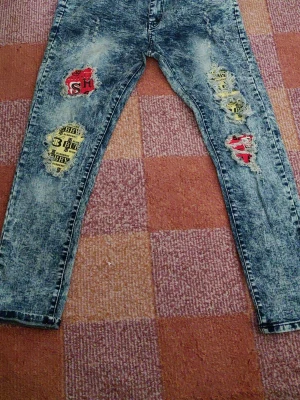 Acid wash jeans med patchdetaljer - Säljer ett par blå acid wash jeans med slitningar och coola patchar i rött och gult med text. Jeansen har smal passform och klassisk femficksdesign. Perfekta för dig som vill sticka ut med en unik streetstyle-look.