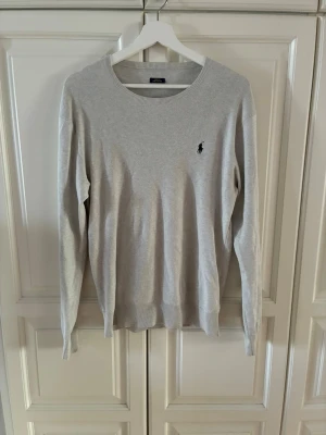 Ljusgrå stickad tröja från Polo Ralph Lauren - Ljusgrå stickad tröja från Polo Ralph Lauren med rund halsringning och diskret broderad logga på bröstet. Tröjan har långa ärmar och ribbade muddar vid ärmslut och nederkant. Perfekt basplagg för en clean och stilren look.