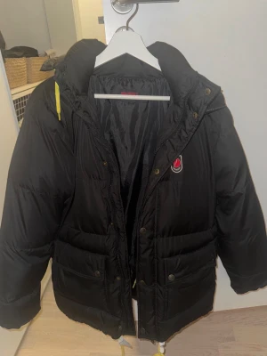 Svart dunjacka från Fjällräven XS - Säljer en svart dunjacka från Fjällräven i storlek XS. Jackan har huva, dragkedja och tryckknappar, samt två stora fickor framtill. Klassisk Fjällräven-logga på bröstet och gul detalj på huvan. Perfekt för kalla vinterdagar.