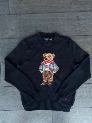 Svart stickad tröja Polo Bear - Svart stickad tröja från Polo Ralph Lauren med ikoniskt Polo Bear-motiv framtill. Rund halsringning och ribbade muddar vid ärmslut och nederkant. Perfekt för dig som gillar klassiska och lekfulla detaljer.