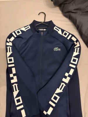 Blå vindjacka från Lacoste Sport - Snygg marinblå vindjacka från Lacoste Sport med vita grafiska detaljer längs ärmarna och klassisk krokodillogga på bröstet. Jackan har dragkedja framtill och hög krage för extra stilpoäng. Perfekt för dig som gillar sportig och modern look.