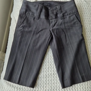 Svart  randiga shorts från Vero Moda - Snygga svarta shorts från Vero Moda med låg midja och vita tunna ränder. Klassisk design med hällor för bälte, fickor fram och bak samt knäppning med dragkedja och knappar. Perfekta för en stilren look.