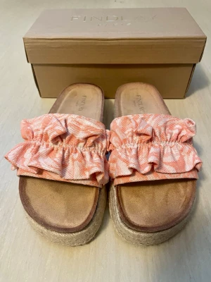 Espandrillos med volang från Findlay Paris i stl 37 - Säljer ett par snygga sandaler från Findlay med breda remmar i orange- och vitmönstrat tyg med volangdetaljer. Sandalerna har en platåsula i flätad jute-look och innersula i mockaimitation. Perfekta för en trendig sommarstil.              Provade ute ett tillfälle, inget att anmärka på.  Stl 37, normal i stl.  Mycket mjuk och skön innersula.  Nypris: 379 sek, Pris nu: 150 sek   