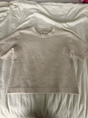 Beige stickad topp från Kappahl - Mysig beige stickad topp från Kappahl med rund halsringning och korta ärmar. Ribbstickade detaljer vid hals, ärmslut och nederkant ger en clean look. Perfekt för dig som gillar enkel och stilren design. Säljer då den aldrig kommer till användning 