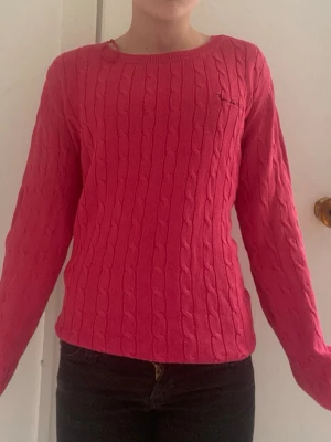 Rosa kabelstickad tröja Gant - Säljer en rosa kabelstickad tröja från Gant med rund halsringning och diskret logga broderad på bröstet. Tröjan har långa ärmar och ribbade muddar vid hals, ärmslut och nederkant. Perfekt för dig som gillar klassisk och stilren design.