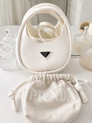 Vit handväska  - Stilren vit handväska i en oval form. Väskan har ett klassiskt emblem i silver framtill och levereras med en matchande dustbag med justerbar axelrem. Perfekt accessoar för en trendig look.