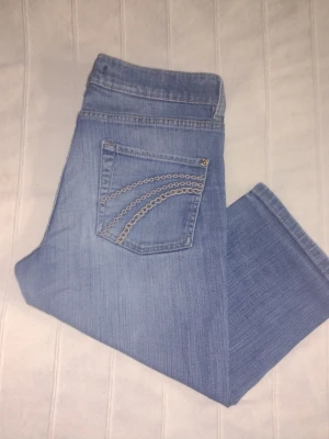 Blå raka jeans/capribyxor med broderade fickor- storlek 42 - Jeans från Lindex i en ljusblå/mellanblå tvätt. Byxorna har en klassisk design med fem fickor - bakfickorna är prydda med dekorativa sömmar. Storlek 42. I bra använt skick.
