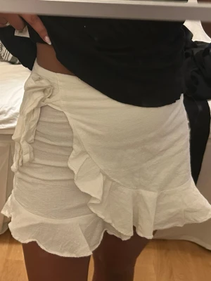 Vit omlottkjol med volang - Supersöt vit kortkjol med omlottdesign och volangkant. Kjolen har en knytning i sidan som ger en extra detalj och är gjord i ett mjukt bomullsliknande material. Perfekt för en fräsch och somrig look!