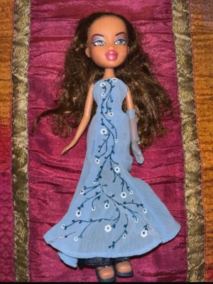 Original Bratz-docka – tidigt 2000-tal Y2K – lång festklänning - Original Bratz-docka från den tidiga Y2K-eran med klassisk Bratz-look och elegant ljusblå långklänning med broderade blommiga detaljer. Dockan har långt brunt hår, markerad makeup och matchande accessoar/handske enligt bilderna.  Dockan är i mycket bra vintage-skick med fint hår, tydligt ansikte och välbevarade kläder. Perfekt för samlare av Bratz, Y2K-leksaker eller nostalgiska mode- och dockentusiaster.  En fin och dekorativ samlardocka från den ikoniska Bratz-eran.  Skick: Mycket bra 