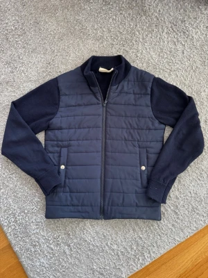 Mörkblå quiltad cardigan från Sayless - Mörkblå cardiganjacka från Sayless med quiltad front och stickade ärmar. Jackan har dragkedja, ståkrage och två fickor med knappstängning. Perfekt för lager-på-lager och en stilren look.