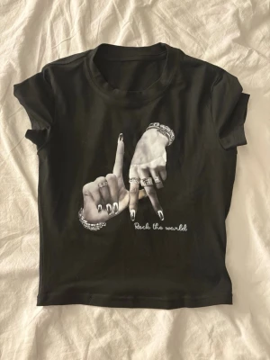 Svart t-shirt med tryck och smycken - Cool svart t-shirt med korta ärmar och tryck av två händer med smycken och texten 'Rock the world' på framsidan. Perfekt för dig som gillar statement-plagg och snygga detaljer. T-shirten har en klassisk rund hals och är lätt att matcha.