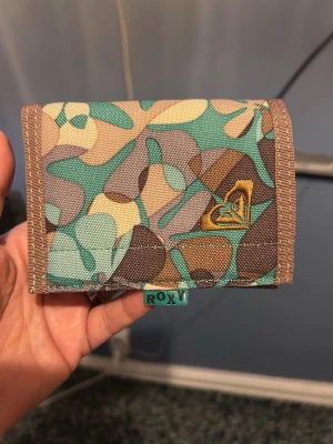 Roxy plånbok med mönster - Plånbok från Roxy i tyg med färgglatt abstrakt mönster i turkos, beige, brunt och grönt. Broderad Roxy-logga i gult på framsidan och bruna kanter. Praktisk och smidig modell med plats för kort och sedlar.