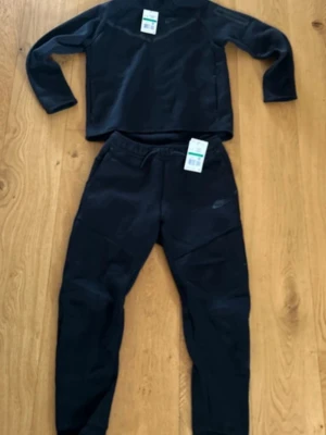 Svart Nike tech set - Mysigt svart loungewear-set från Nike med långärmad tröja och matchande byxor. Setet har en enkel och sportig design med diskret Nike-logga på både tröja och byxa. Perfekt för chill hemma eller avslappnade dagar. Mjukt material och bekväm passform. STORLEK XL I BARNSTORLEK