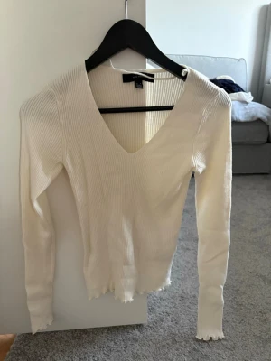 Vit ribbad v-ringad topp - Vit långärmad topp från Vero Moda med ribbad struktur och v-ringning. Modellen är figurnära och har vågiga kanter vid ärmslut och nederkant. Perfekt att styla med jeans eller kjol för en trendig look. Använd fåtal gånger