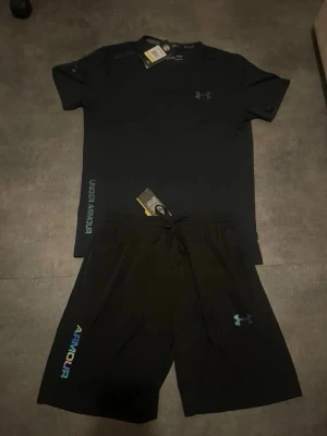 Svart Under Armour - Snyggt svart träningsset från Under Armour med t-shirt och shorts. T-shirten har tryck i neonfärger och logga på bröstet, medan shortsen har elastisk midja och färgglada detaljer längs sidan. Perfekt för gymmet eller löpning. Materialet är lätt och andas.