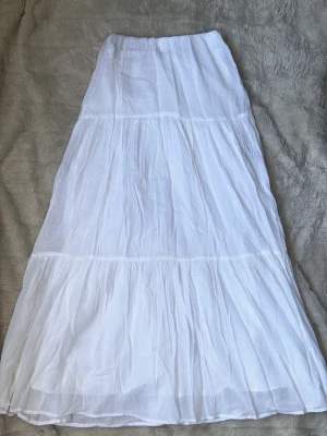 Vit långkjol i bomull - Säljer en luftig och flowy vit långkjol i bomull. Kjolen har flera lager och en lätt rynkad design som ger en bohemisk känsla. Perfekt för varma dagar och enkel att matcha med olika toppar. Strl XS🤍 Har bara legat i en garderob och aldrig kommit till användning. Resår i midjan 