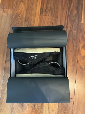 Svarta Saint Laurent sneakers - Svarta sneakers från Saint Laurent med vit sula och diskret logga på sidan. Skorna har klassisk låg profil, snörning och är tillverkade i slitstarkt tygmaterial. Perfekta för dig som gillar stilrena och tidlösa sneakers med exklusiv känsla. Storlek 42 men passar 41! Skriv gärna om ni har funderingar!