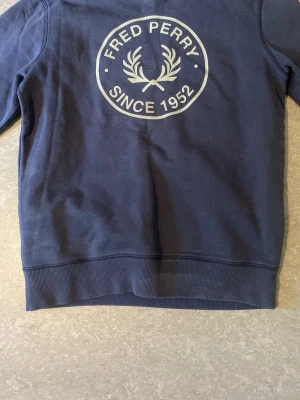 Blå Fred Perry hoodie med tryck - Mörkblå hoodie från Fred Perry med stort vitt tryck på ryggen där det står 'Fred Perry Since 1952' och den klassiska lagerkransen. Tröjan har ribbade muddar och är tillverkad i mjukt bomullsmaterial. Perfekt för en avslappnad och stilren look.