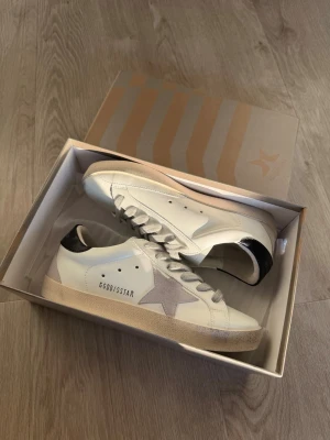 Vita Golden Goose sneakers med stjärna - Snygga vita sneakers från Golden Goose med grå stjärndetalj på sidan och svart hälparti. Skorna har klassisk låg modell, vita skosnören och beige sula. Perfekta för dig som gillar en stilren men trendig look.