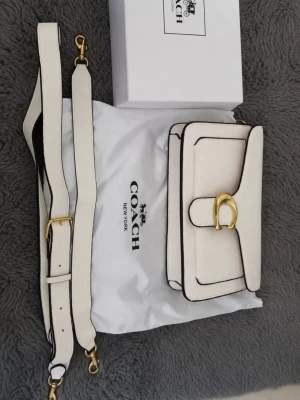Vit Coach tabby bag 26  - Snygg vit handväska från Coach med justerbar axelrem och guldfärgat spänne framtill. Väskan har svarta kanter som ger en cool kontrast och klassisk look. Perfekt för dig som gillar stilrena accessoarer med exklusiv känsla.