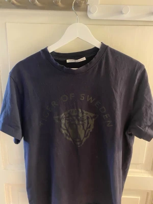 Mörkblå Tiger of Sweden t-shirt - Snygg mörkblå t-shirt från Tiger of Sweden med diskret tryck av tigerhuvud och text på bröstet. Klassisk rund hals och korta ärmar. Tillverkad i mjuk bomull som känns skön mot huden. Perfekt för en clean och stilren look.