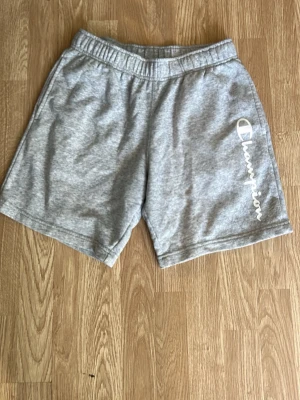 Grå shorts från Champion - Snygga grå shorts från Champion med elastisk midja och logga tryckt i vitt längs ena benet. Tillverkade i mjuk bomullsmix som passar perfekt för chill eller träning. Klassisk passform och enkel design.