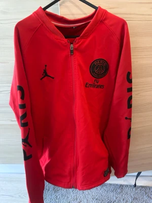 Röd PSG x Jordan ziptröja - Röd ziptröja från Jordan i samarbete med Paris Saint-Germain. Tröjan har svarta tryck med PSG-logga, Fly Emirates och Jordan-logga på bröstet samt texten PARIS längs ärmarna. Perfekt för dig som gillar fotboll och streetwear.