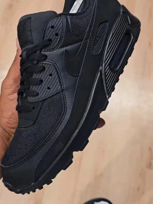 Nike Air Max 90 Triple Black sneakers - Snygga Nike Air Max 90 i helsvart färg. Skorna har en klassisk siluett med mesh och läderdetaljer, samt den ikoniska Air Max-dämpningen i sulan. Perfekta för dig som gillar en clean och stilren look med mycket komfort.