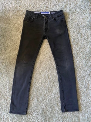 Jacob Cohën Bard strl 32 svart/grå - Säljer ett par svart/grå jeans från Jacob Cohen i modellen Bard. Jeansen hqr en smal och är tillverkade i stretchigt denimtyg för extra komfort. Snygga detaljer som svarta sömmar och fet patch där bak. Mycket bra skick och inga defekter. Mått: midja ~42 cm och längd ~106 cm. Hör av er vid frågor och funderingar!