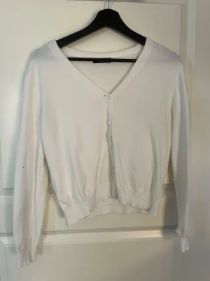 Vit kofta med v-ringning - Klassisk vit kofta med v-ringning och knäppning framtill. Långärmad modell med ribbade muddar och nederkant. Perfekt att slänga över en topp eller klänning för en clean look. Aldrig anvönd.