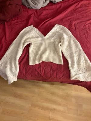 Vit stickad tröja med v-ringning - Snygg vit stickad tröja med djup v-ringning både fram och bak. Tröjan har vida ärmar med hålmönster och ribbstickad midja. Perfekt för dig som gillar en trendig och feminin look. Säljer för att den är lite liten