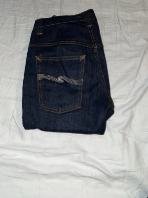 Mörkblå jeans från Nudie Jeans - Säljer ett par mörkblå jeans från Nudie Jeans med klassisk femficksdesign och kontrastsömmar. Jeansen har en smal passform och är tillverkade i denim med snygg broderad bakficka. Perfekta för dig som gillar stilrena och tidlösa jeans.