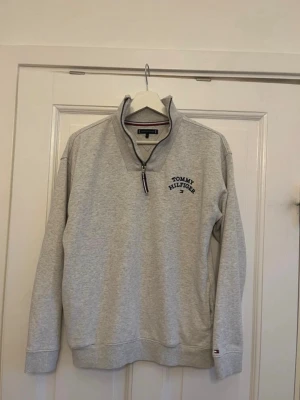 Grå halvzip från Tommy Hilfiger - Snygg grå halvzip-tröja från Tommy Hilfiger med broderad logga på bröstet och klassiska Hilfiger-detaljer vid dragkedjan och ärmslutet. Tröjan har hög krage och ribbade muddar. Perfekt för en avslappnad och stilren look.