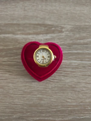 Guldfärgad ring med klocka - Unik guldfärgad ring med inbyggd klocka och vit urtavla med romerska siffror. Ringen har en klassisk rund form och detaljer i svart på urtavlan. Perfekt accessoar för dig som vill sticka ut och kombinera stil med funktion.