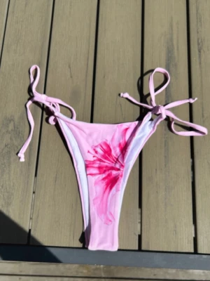 Rosa bikiniunderdel med blomma - Säljer en ljusrosa bikiniunderdel med knyt i sidorna och ett snyggt blommigt mönster i rosa och rött på ena sidan. Perfekt för sommaren och strandhäng! Helt ny