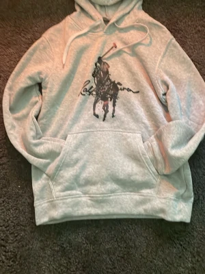 Grå hoodie från Ralph Lauren - Snygg grå hoodie från Ralph Lauren med stor svart polospelare tryckt på bröstet och klassisk känguruficka. Mjuk bomullsblandning och justerbar huva med vita snören. Perfekt för en avslappnad och trendig look.