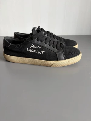 Svarta Saint Laurent sneakers - Svarta låga sneakers från Saint Laurent med handskriven logga på sidan. Skorna har en ovandel i canvas och detaljer i läder, samt en beige sula som ger en vintagekänsla. Klassisk snörning och slitna detaljer för en cool, avslappnad stil.