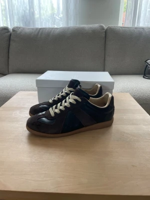 Maison Margiela Gats - Ett par Maison Margiela Gats i en brun och svart färg. Skorna är i bra skick på utsidan, men dem har varsitt litet hål inuti skorna, det varken känner man eller märker man när man har dem på sig. Utöver det är dem i mycket bra skick, lädret är väl omhändertaget. Box, dustbag och visitkort tillkommer. Skriv om du har någon fråga! Priset kan diskuteras!