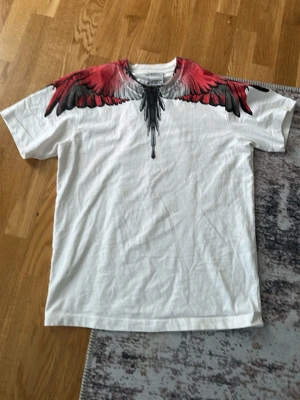 Vit t-shirt med röda vingar från Marcelo Burlon - Säljer en vit t-shirt från Marcelo Burlon med ikoniskt tryck av röda och svarta vingar över axlarna och ner mot bröstet. Klassisk passform och rund hals. Perfekt statement-plagg för dig som vill sticka ut.