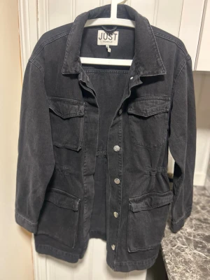 Svart jeansjacka från Just Female - Overshirt i svart denim från Just Female med fyra stora fickor framtill och silverfärgade knappar. Rak och avslappnad passform med klassisk krage. Perfekt lager-på-lager-plagg för en trendig look. Storlek M