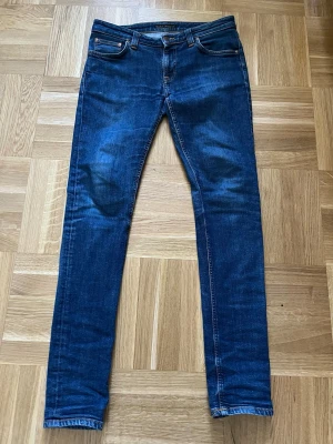 Blå jeans från Nudie Jeans, W31 L34 - Säljer ett par klassiska blå jeans från Nudie Jeans i storlek W31 L34. Jeansen har smal passform, snygga slitningar och kontrastsömmar. Tillverkade i denim med två bakfickor och Nudie Jeans logga på fickorna. Perfekta för en avslappnad och trendig look.