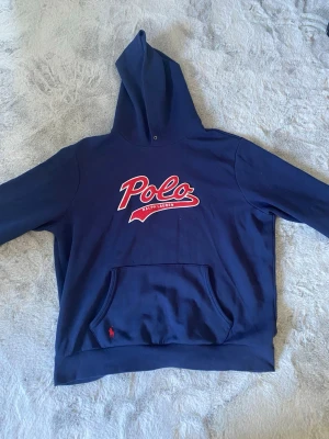 Blå hoodie från Polo Ralph Lauren - Snygg mörkblå hoodie från Polo Ralph Lauren med stor röd och vit logotyp på bröstet. Hoodien har en klassisk huva och en stor magficka framtill. Perfekt för dig som gillar streetstyle och vill ha något bekvämt och trendigt. XL men passar L och M. DM för frågor!